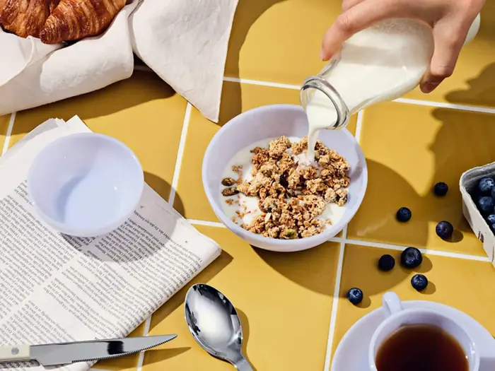 En hånd hælder mælk i en skål med granola på et bord med gule fliser, med en ske, en avis, blåbær, te og en croissant i nærheden.