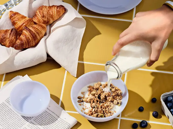 En hånd hælder mælk i en skål med granola på et gult kakkelbord med croissanter, blåbær, en avis og tomme skåle i nærheden.
