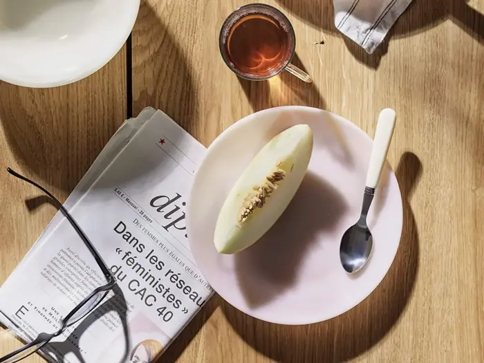 Et bord med en avis, glas, en skål, et glas te og en tallerken med en melonskive og en ske i skarpt sollys.