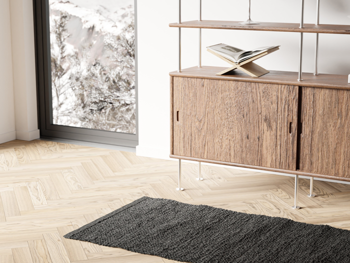 Et træskab med metalben står ved et stort vindue; en åben bog hviler ovenpå, og et mørkt tekstureret tæppe ligger på et lyst sildebensgulv. Sneklædte træer er synlige udenfor.