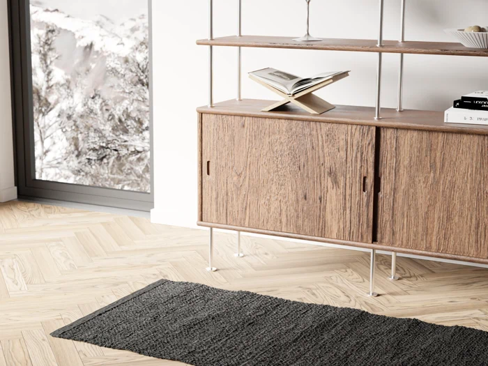 Et træskab med hylder og metalben står på et lyst trægulv ved siden af et vindue, der viser sneklædte bjerge; et gråt tæppe ligger foran. En åben bog og en skål står på hylderne.