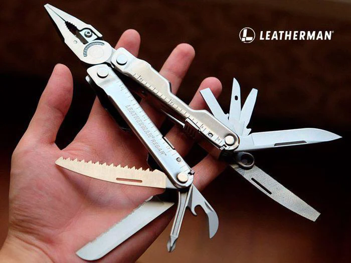 En hånd holder et åbent Leatherman multiværktøj, der viser tænger, knive, sav, file, skruetrækkere og andet værktøj. Leatherman-logoet vises i øverste højre hjørne.