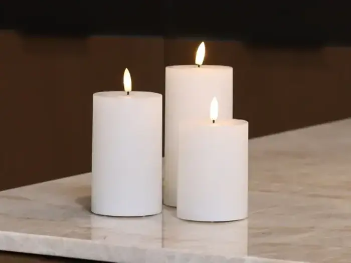 Tre hvide bloklys i forskellige højder tændes og placeres på en marmoroverflade, der kaster et varmt skær mod en mørk baggrund.