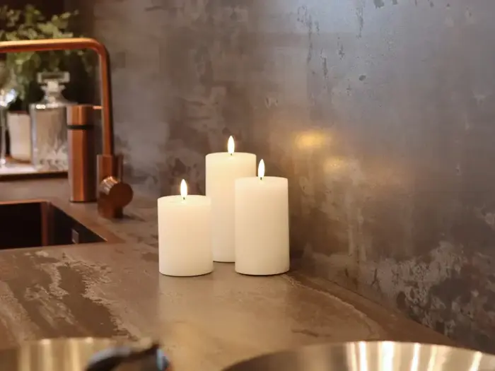 Tre hvide bloklys er tændt og placeret på et køkkenbord i nærheden af en kobbervask og vandhane, hvilket skaber en hyggelig og varm atmosfære.