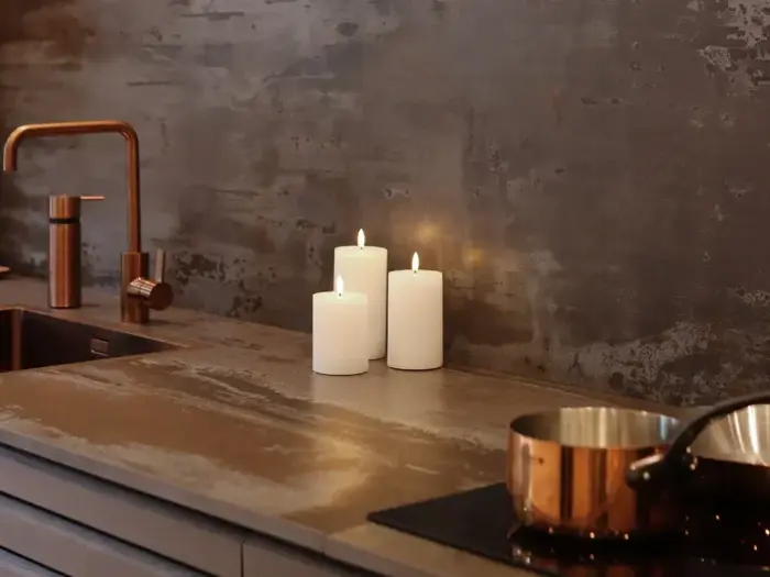 Moderne køkken med en metallisk bordplade, tre tændte hvide bloklys, en kobbervask og vandhane samt en kobbergryde på komfuret.