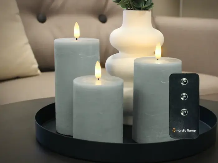 Tre grå bloklys med realistiske flammer er placeret på en sort bakke ved siden af en hvid vase. En fjernbetjening med knapper er synlig i forgrunden.