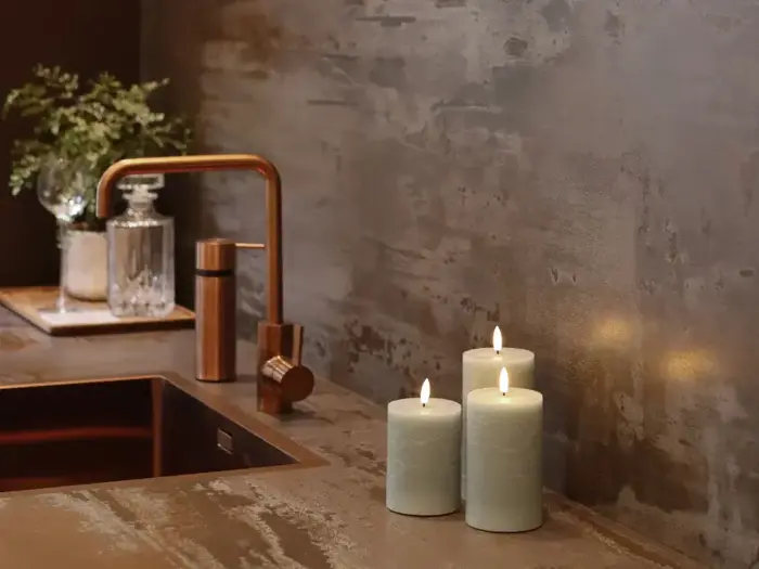 Tre tændte bloklys står på et moderne badeværelsesbord ved siden af en kobbervandhane med en sæbedispenser i glas og en lille vase med grønne planter i baggrunden.