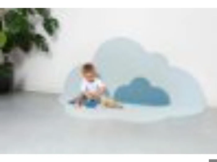 Et lille barn sidder på gulvet foran en stor, dekorativ sky, der er skåret ud mod en hvid væg med en potteplante i baggrunden.