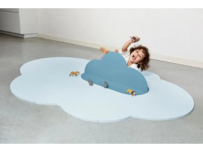 Et smilende barn leger med legetøjsdyr på en stor, lyseblå skyformet måtte med en blå skyformet skillevæg i midten, indendørs på et gråt gulv.