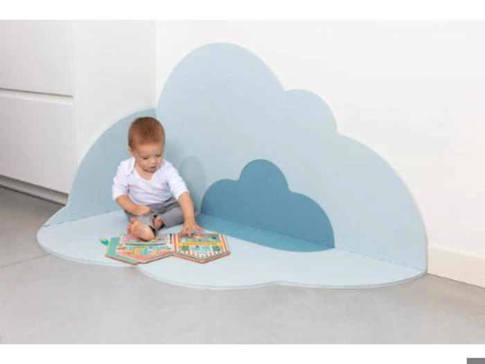 En baby sidder på en skyformet blå måtte i hjørnet og leger med en åben bog eller et stykke legetøj på gulvet.