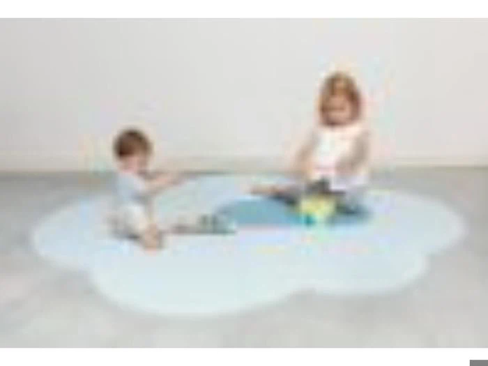 To små børn sidder og leger på en skyformet blå måtte i et lyst, minimalistisk rum med et lysegråt gulv og en almindelig hvid væg.