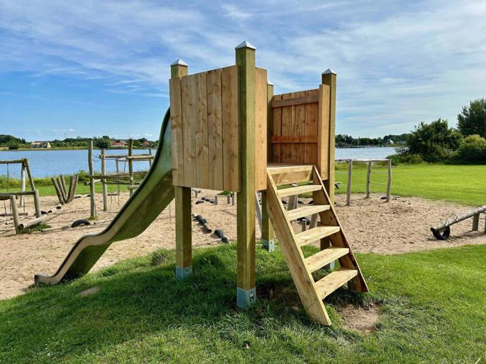 En legeplads i træ med trapper og en grøn rutsjebane står på en græsplæne nær et sandområde med en sø og træer i baggrunden under en blå himmel.