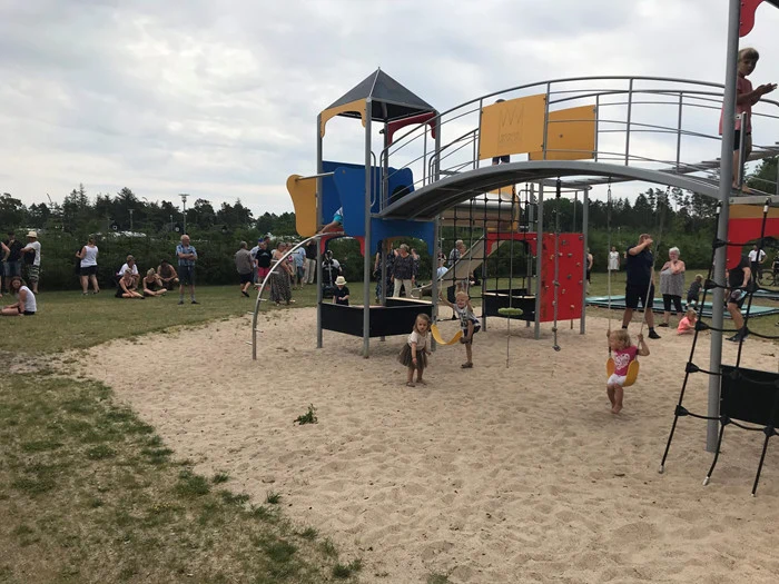 Børn leger på gynger og klatrestativer på en legeplads, mens voksne og andre børn samles på græsset i baggrunden under en overskyet himmel.