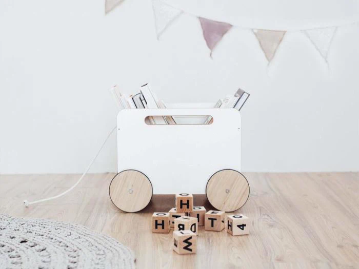 En hvid legetøjsvogn af træ med bøger i står på et lyst trægulv, træklodser med bogstaver ligger spredt foran, og pastelfarvede vimpler hænger på væggen bagved.