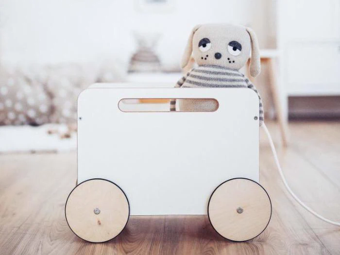 En blød, stribet tøjhund sidder i en hvid legetøjsvogn af træ med store runde hjul på et trægulv i et lyst rum.