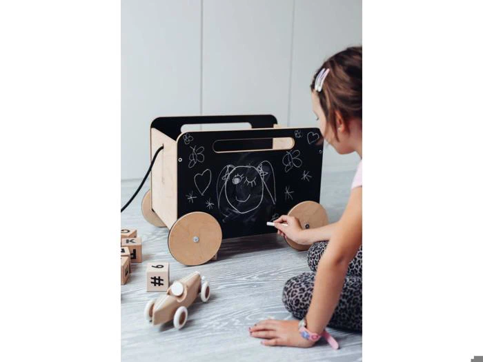 En ung pige tegner et ansigt med kridt på en tavle på en legetøjsvogn af træ med klodser og en legetøjsbil på gulvet i nærheden.