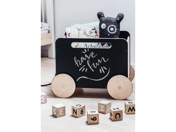 En legetøjsvogn med træhjul rummer bøger og en sort plysbjørn. Have fun står der med kridt på siden. Bogstavklodser af træ ligger spredt på det lyse gulv i nærheden.
