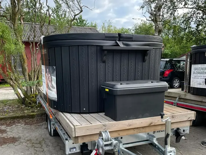 Et stort sort spabad står på en trævogn ved siden af en sort opbevaringsboks med træer, en rød bygning og parkerede biler i baggrunden.