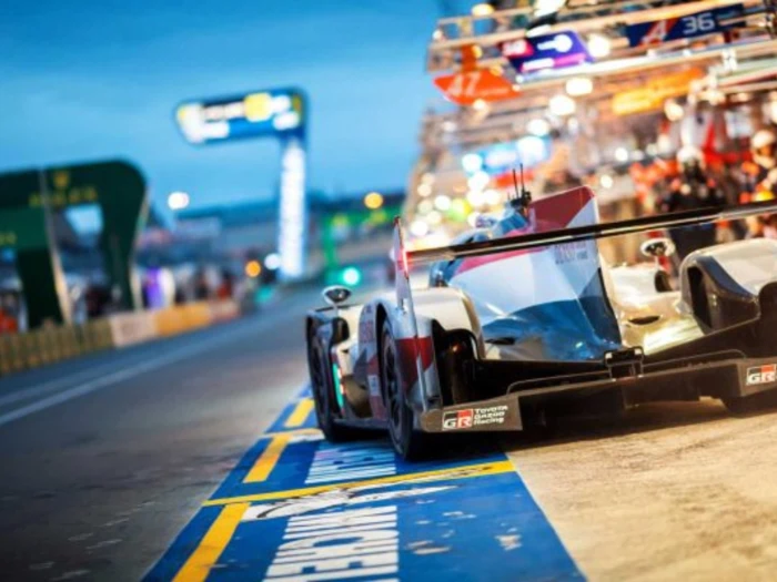 En racerbil kører ind i en stærkt oplyst pitbane i skumringen med teammedlemmer og skilte synlige i baggrunden.