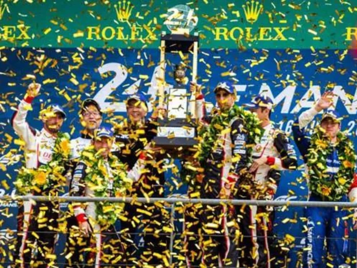 Racerkørere på et podium fejrer en sejr med konfetti, der falder ned, og de holder et stort trofæ og bærer grønne laurbærkranse, med et 24 og Rolex-banner i baggrunden.