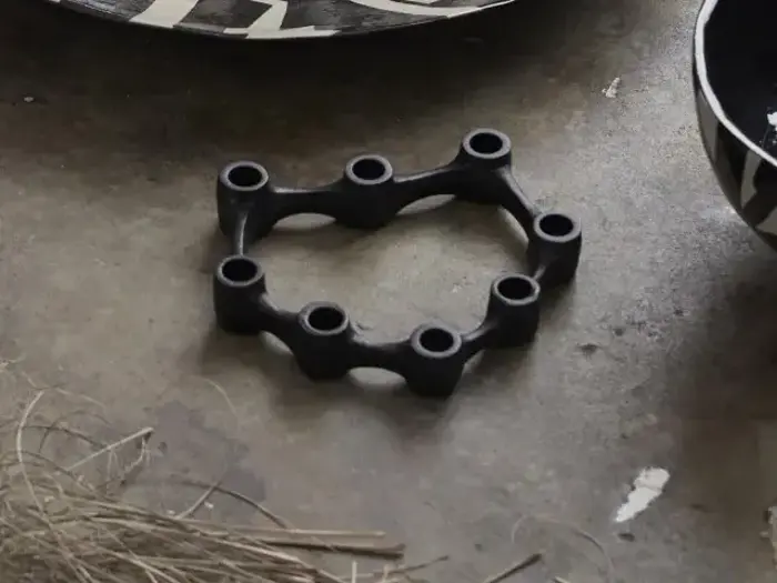 Et sort, sekskantet objekt med seks runde åbninger hviler på en betonflade ved siden af mønstrede skåle og spredte kviste.