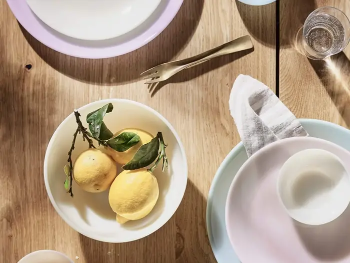 En bordscene med en skål med citroner og blade, pastelfarvede tallerkener, en guldgaffel, et glas vand og en serviet på en lys træoverflade.