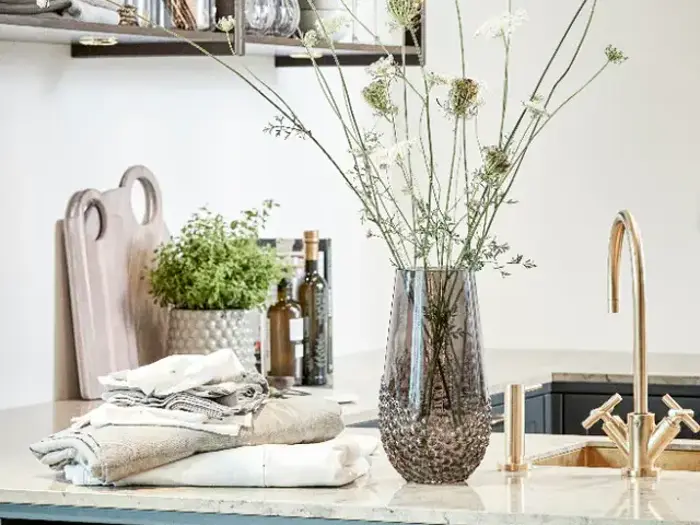 Et køkkenbord med en glasvase med høje hvide blomster, foldede håndklæder, krydderurter i potter, skærebrætter og flasker ved siden af en guldvandhane.