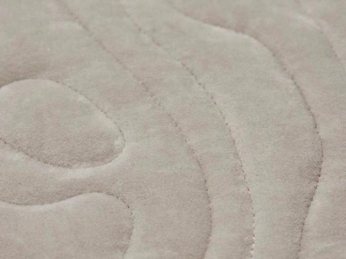 Nærbillede af et beige, struktureret stof med bløde, bølgede syninger, der ligner quiltet eller polstret betræk.