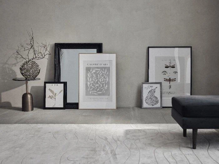 Minimalistisk rum med indrammede botanik- og insektprint, der læner sig op ad en grå væg, en tørret gren i en vase og en mørk bænk på et mønstret tæppe.