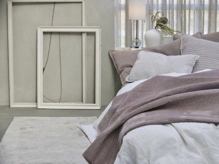 En hyggelig seng med bløde puder og et lilla tæppe ved siden af to tomme, hvide billedrammer, der læner sig op ad en væg i et fredfyldt, blødt oplyst soveværelse.