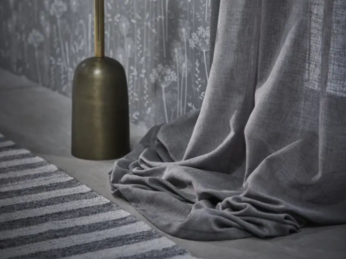 Et gråt gardin er trukket ned på gulvet ved siden af en messingfarvet lampe og et grå- og hvidstribet tæppe med blomstermønstret tapet i baggrunden.