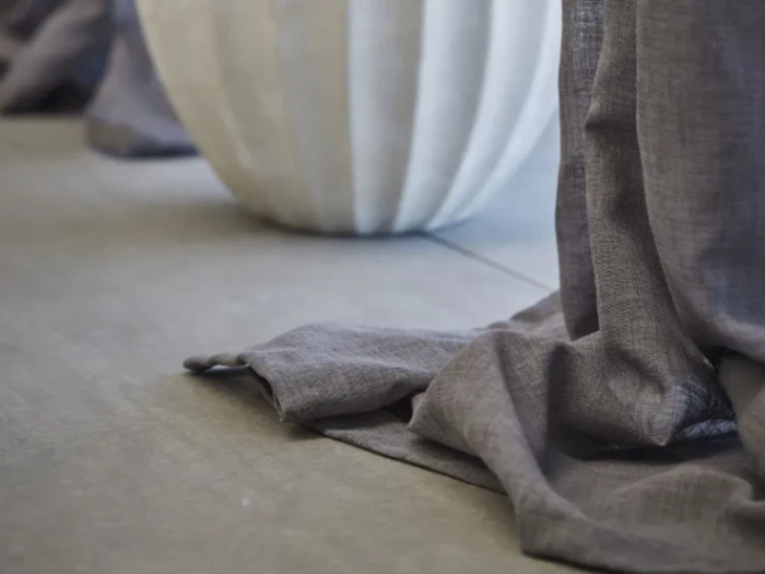 Nærbillede af grå stofgardiner, der flyder sammen på et gråt klinkegulv, med en stor hvid tekstureret vase i baggrunden.