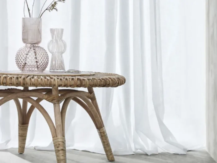 Et flettet bord med to glasvaser står foran hvide, gennemsigtige gardiner, og et blødt, naturligt lys trænger igennem.