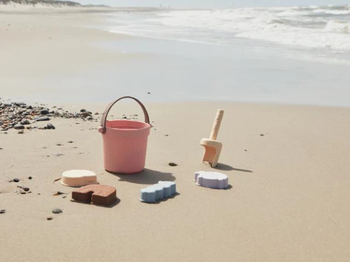 En lyserød spand, en legetøjsskovl og farverige sandforme står på sandstranden nær kysten med bølger i baggrunden.