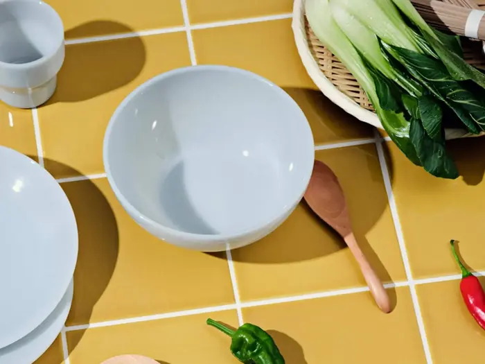 En hvid skål, en træske, grøntsager og tallerkener er anbragt på en gul flise.
