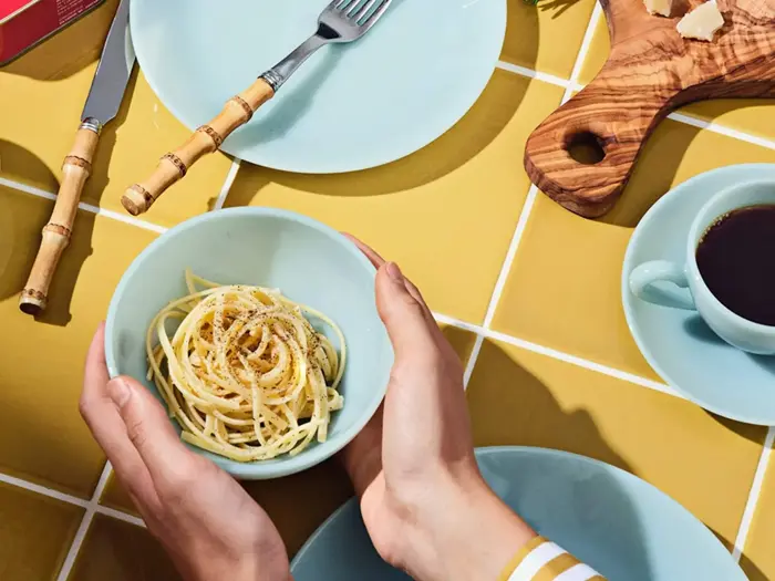 Hænderne holder en skål spaghetti over et gult flisebord med tallerkener, bestik, en kop kaffe og et træbræt med ost.