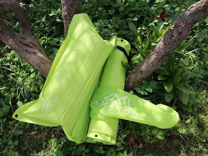 To lysegrønne AMAROOQ-campingsoveunderlag rulles ud og ind og hviler mod et træ blandt frodige grønne planter.