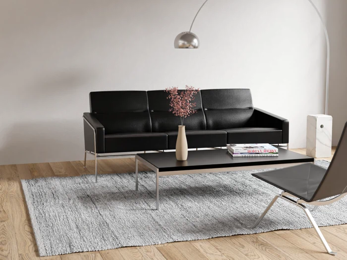 En moderne stue med en sort sofa, en matchende stol, et sort sofabord med bøger og en vase, et gråt tæppe og en gulvlampe, alt sammen på et lyst trægulv.