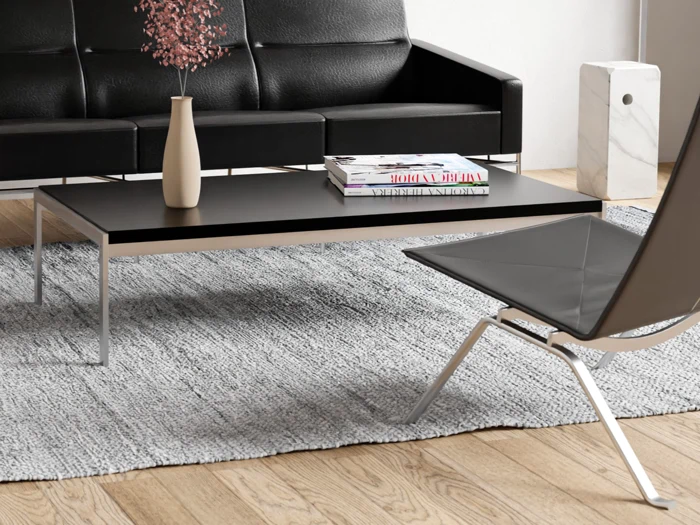 En moderne stue med en sort sofa, et sort sofabord med en beige vase og magasiner, en grå stol og et struktureret gråt tæppe på et trægulv.