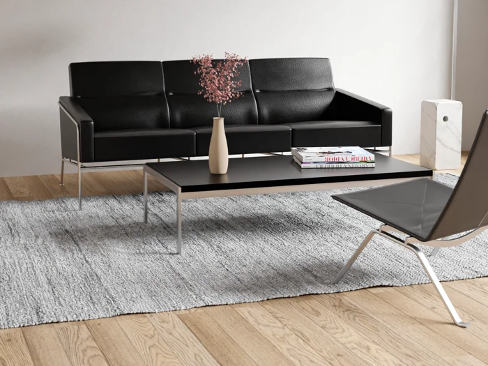 En moderne stue med en sort lædersofa, et sort sofabord med magasiner og en beige vase, en grå stol og et gråt tæppe på et trægulv.