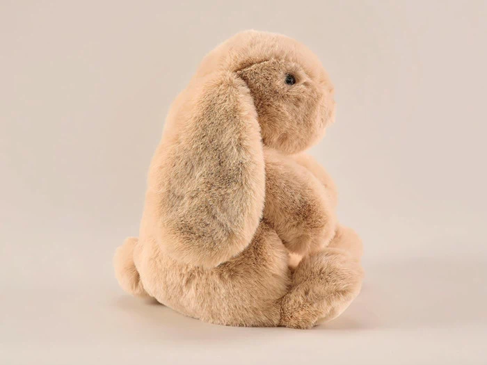 En blød, lysebrun kanin i plys med hængeører sidder oprejst på en almindelig, lys baggrund.