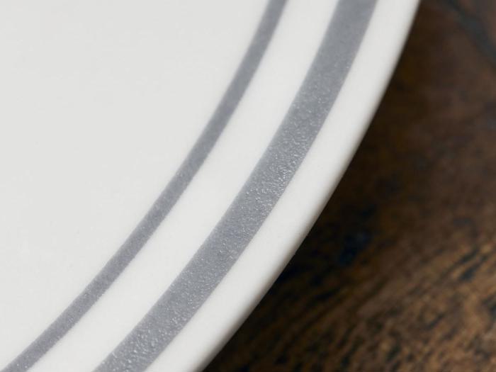 Nærbillede af en hvid tallerken med to grå striber nær kanten, placeret på en brun træoverflade.