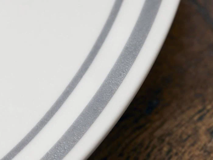 Nærbillede af en hvid tallerken med to grå striber nær kanten, placeret på en brun træoverflade.