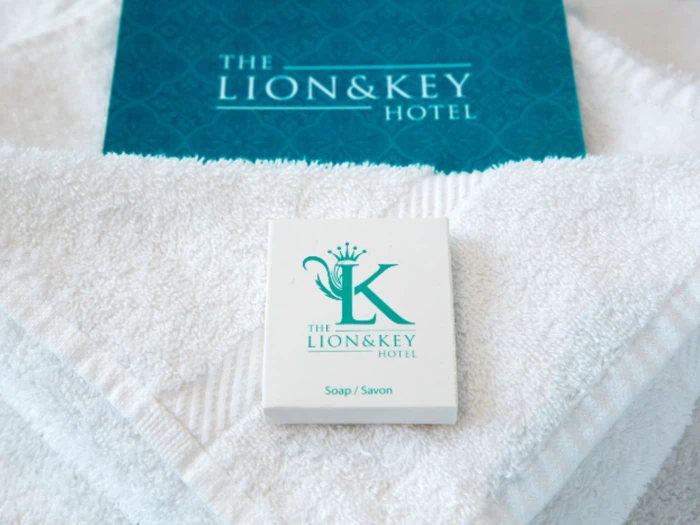 Et hvidt stykke sæbe og en blå brochure, begge med påskriften The Lion & Key Hotel, er placeret på sirligt foldede hvide håndklæder.