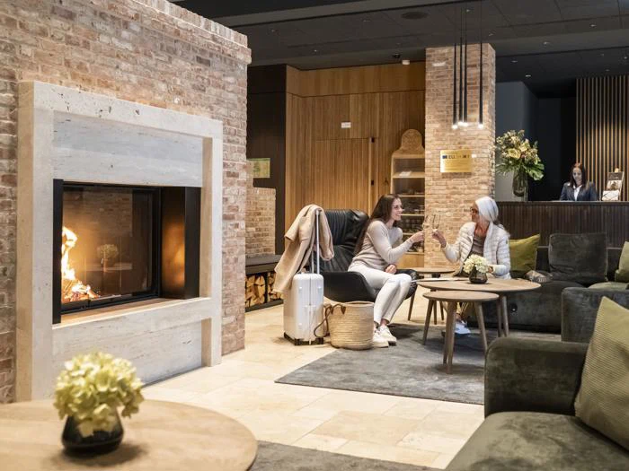 To kvinder sidder og snakker i en hyggelig hotellobby med pejs, bagage og varm indretning; en receptionist står ved skrivebordet i baggrunden.