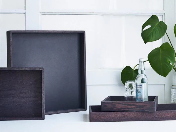 Tre mørke træbakker er stablet og arrangeret på en hvid flade ved siden af en grøn potteplante, med et glas og en klar flaske placeret på den mindste bakke.