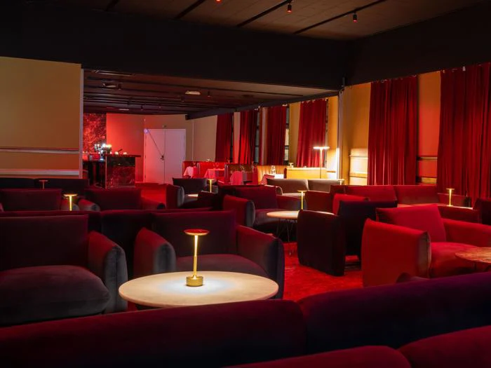 En svagt oplyst lounge med røde lænestole, runde borde og små bordlamper, røde gardiner på væggene og en bar i baggrunden.