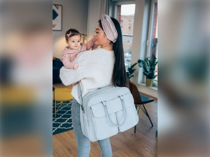 En kvinde med en baby på hoften, begge iført pastelfarvede pandebånd, står indendørs med en stor lysegrå taske over skulderen.