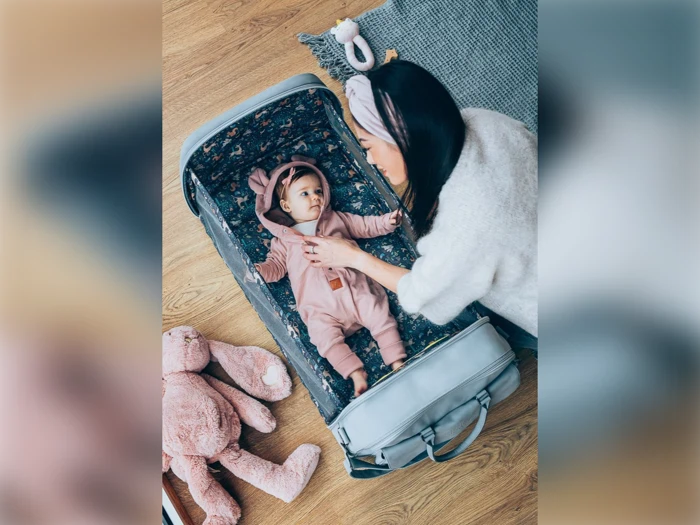 En kvinde kigger kærligt på en baby, der ligger i en kuffertvugge. Babyen har en lyserød sparkedragt på med bjørneører. En udstoppet lyserød kanin ligger i nærheden på gulvet.