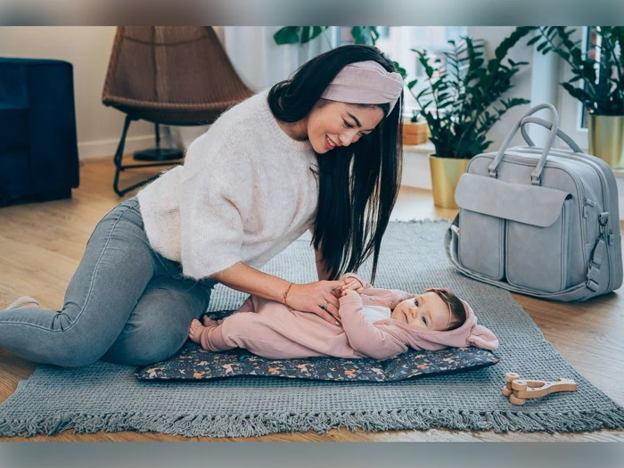 En smilende kvinde knæler på et tæppe indendørs og leger med en baby i lyserødt tøj, der ligger på en måtte; en grå pusletaske står i nærheden.